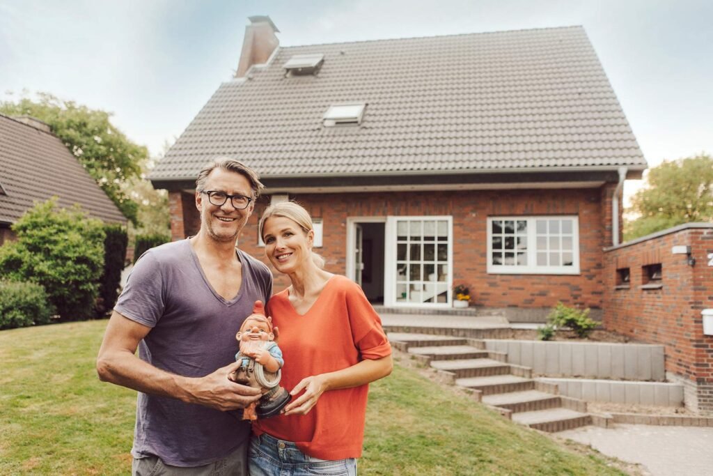 Pareja sonriente posando para retrato con un duende de jardín en sus manos, de fondo la fachada de casa y arbolada.